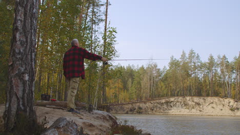 angling-at-weekend-middle-aged-fisherman-is-standing-on-coast-of-forest-river-under-high-trees-and-relaxing
