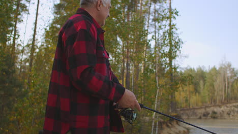 adult-fisherman-is-rotating-reel-of-rod-during-spin-fishing-in-freshwater-standing-on-shore-of-river-in-forest-at-autumn-day