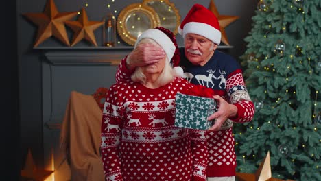 Senior-grandfather-covering-eyes-of-old-grandmother-with-surprise-Christmas-gift-present-box-at-home