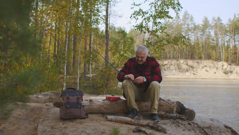 grey-haired-fisherman-is-putting-bait-on-hook-of-rod-preparing-to-spin-fishing-on-shore-of-river-in-forest-camping-and-hiking