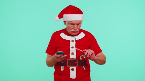 Älterer-Weihnachtsgroßvater-Mit-Kreditkarte-Und-Smartphone,-Der-Geld-Beim-Online-Einkauf-überweist