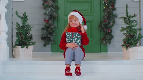 Freudig-Lächelndes-Kleinkind-Sitzt-Auf-Der-Dekorierten-Veranda-Des-Hauses-Und-Hält-Eine-Weihnachtsschachtel-In-Der-Hand