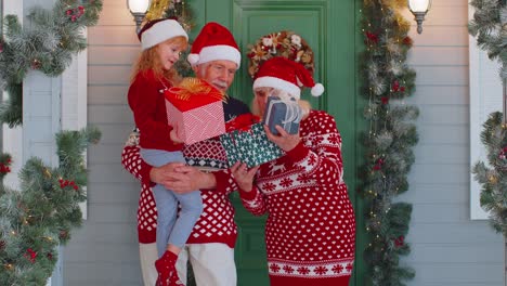 Happy-senior-grandparents-with-granddaughter-holding-many-gift-boxes-near-Christmas-house,-celebrate