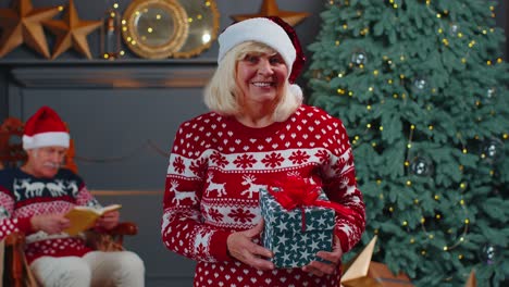 Ältere-Großmutter-Im-Festlichen-Pullover-Präsentiert-Weihnachtsgeschenkbox,-Lächelt-Und-Blickt-In-Die-Kamera