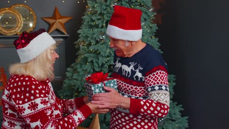 Senior-grandmother-presenting-Christmas-gift-box-to-surprised-grandfather-at-decorated-home-room
