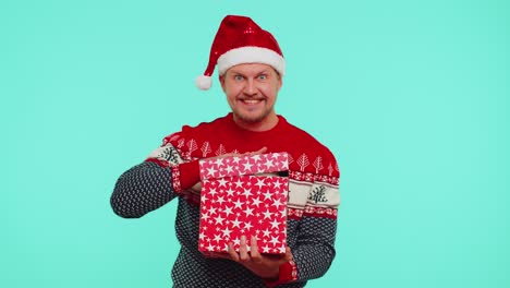 Funny-30s-years-old-man-wears-red-New-Year-sweater-hat-unwrapping-gift,-opening-box-with-pet-cat