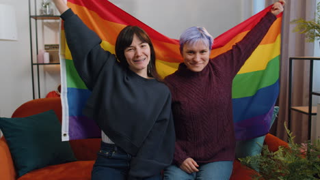 Two-lesbian-women-family-couple-or-girl-friends-holding-LGBT-people-gay-pride-flag-in-hands-at-home