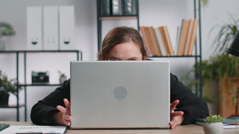 Confident-young-business-woman-hiding-behind-laptop-computer,-looking-at-camera,-spying,-peeping