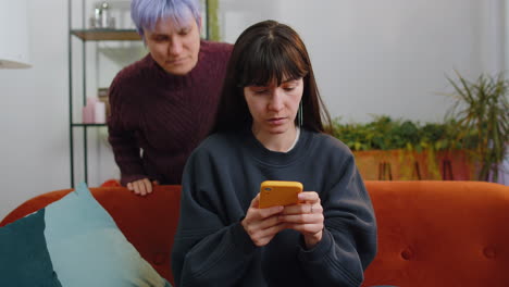 Two-young-lesbian-women-family-married-couple-working-on-smartphone,-shopping-at-home-living-room