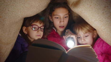 Children-girl-kids-under-blanket-reading-interesting-fairytale-story-book-using-flashlight-at-home