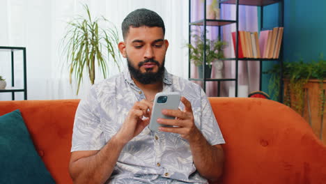 Indian-Arabian-young-man-sitting-on-sofa-using-smartphone-share-messages-on-social-media-application