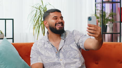 Happy-Indian-man-taking-selfie-on-smartphone-communicating-video-call-home-online-with-subscribers