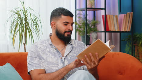 Indian-man-reading-interesting-book-turning-pages-smiling-enjoying-literature-taking-a-rest-on-sofa