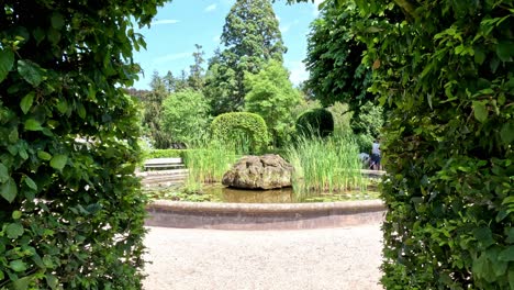 Walking-Through-a-Garden-Gate-Made-From-a-Round-Topped-Hedge-Towards-a-Small-Fountain-Covert-with-Water-Lilies-at-the-Botanical-Garden-in-Karlsruhe