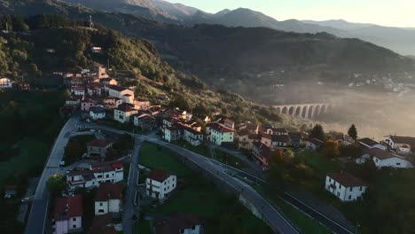 small-italian-typical-town-with-mist-at-golden-hour-in-morning,-aerial-drone-view,-hill-and-mountain-in-background,-with-sun-flares,-tuscany,-italy,-europe