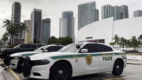 Wide-shot-of-Miami-Dade-Police-Department-car-parked-with-civil-defense-siren-on,-Urban-skyline-buildings-in-the-background