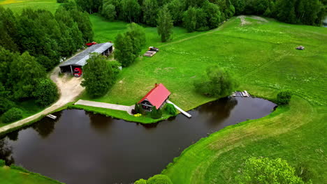 Red-Roof-Farm-House-on-Rural-Farmland,-Lake-House,-Surrounded-by-Forest,-Lush-Green-Grass,-Aerial-Drone-Establishing-Shot,-Wide-Angle