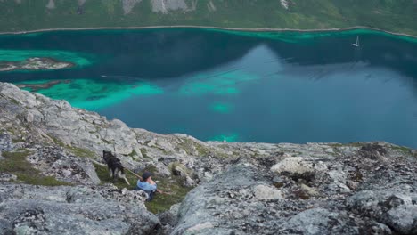 Female-Wanderlust-Sitting-On-Rocky-Hill-Salberget-With-Pet-Dog-Overlooking-Calm-Blue-Lake-In-Norway