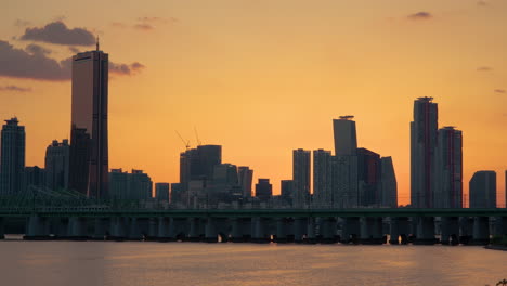 Zug-überquert-Den-Han-Fluss-Und-Fährt-Auf-Der-Eisenbahnbrücke,-Skyline-Der-Insel-Seoul-Yeouido-Mit-63-Gebäuden-In-Der-Silhouette-Während-Des-Malerischen-Orangefarbenen-Sonnenuntergangs,-Südkorea