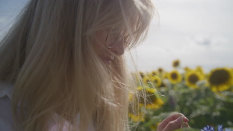 Blonde-woman-smells-purple-flowers-by-sunflower-field,-side-close-up
