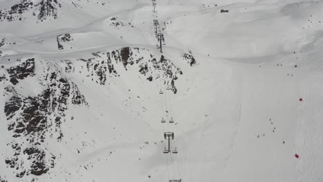 Tilt-down-drone-shot-of-very-long-chairlift,-huge-mountain-range,-ski-slopes-and-skiers-riding-single-file-snake-run