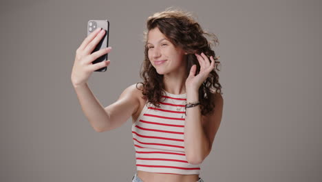 Brunette-girl-fixes-hair-and-takes-selfie-with-phone,-studio-shot