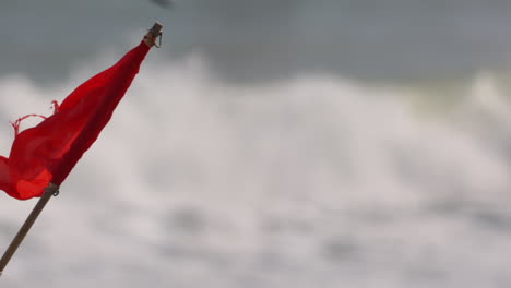 Tight-shot-of-red-flag-in-the-ocean-wind,-with-waves-crashing-in-the-background