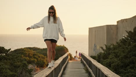 Young-woman-having-fun-walking-along-a-wooden-railing-at-sunset