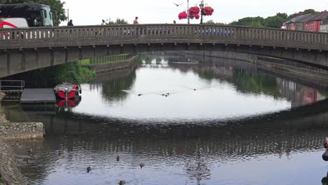 Tourist-boat-docking-near-bridge-in-Kilkenny-City-centre-with-bus-and-people-crossing-the-bridge-in-summer