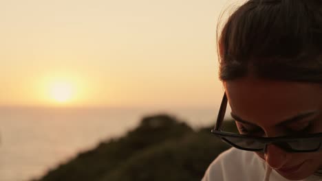 Portrait-of-thoughtful-young-woman-with-sunglasses-looking-down-with-sun-setting-in-background