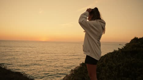 Young-girl-in-sweatshirt-touching-hair-on-sea-cliffs-at-sunset