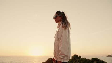 Attraktive-Frau-Mit-Sonnenbrille,-Die-Bei-Sonnenuntergang-Auf-Den-Küstenklippen-In-Die-Kamera-Blickt