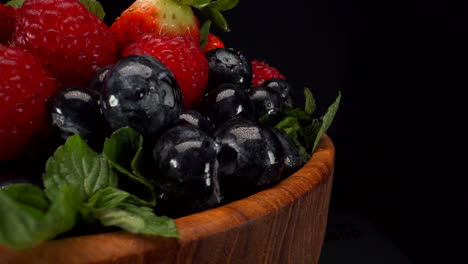 Close-up-rotating-forest-berries-in-a-wooden-bowl-with-black-background,-strawberries,-blueberries,-raspberries,-healthy-fruits,-4K-static-shot
