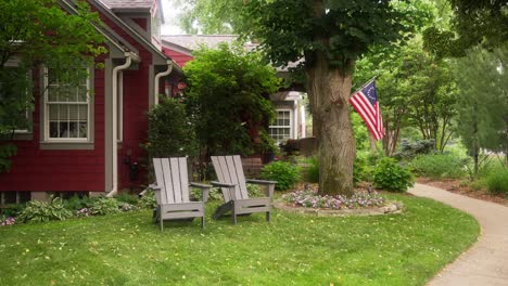 Statische-Aufnahme-Von-Zwei-Adirondack-Stühlen-Im-Vorgarten-Eines-Roten-Hauses-Mit-Einer-Amerikanischen-Betsy-Ross-Flagge-Im-Hintergrund