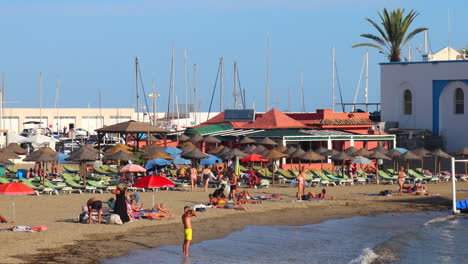 People-enjoying-a-lovely-sunny-beach-day-in-Marbella-port,-fun-sea-vacation-holiday-destination-in-Malaga-Spain,-4K-static-shot
