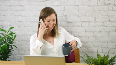 Young-female-business-woman-taking-a-break-chatting-on-phone-and-drinking-coffee-in-an-office-sitting-at-a-desk