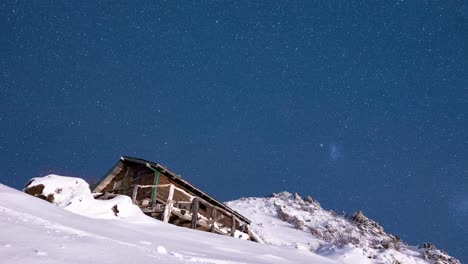 Celestial-night-starry-sky-orbits-high-above-wooden-cabin-on-top-of-snowy-mountain-peak,-time-lapse