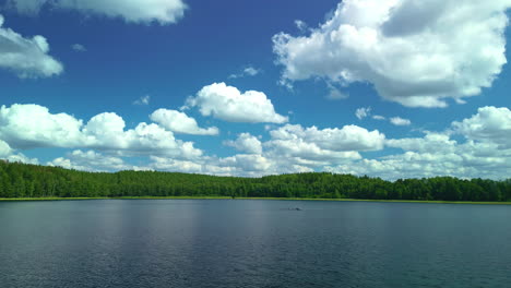 Nubes-Blancas-En-Un-Cielo-Azul-Sobre-Un-Vasto-Lago-Con-Bosques-En-La-Orilla