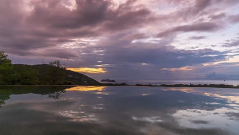 Storm-grey-surge-into-sky-covering-bright-red-yellow-fluffy-clouds,-reflection-in-calm-water-below,-time-lapse