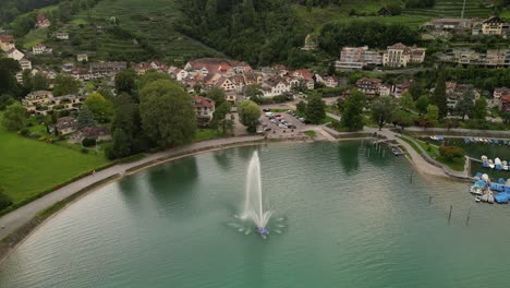 bird-view-flying-towards-a-fountain-splashing-water-in-air-placed-inside-a-lake-cars-parked-along-the-shore-and-brown-houses-standing-under-the-hills-beautiful-architectures-peaceful-environment