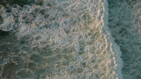 Top-down-aerial-shot-over-white-foaming-waves-in-a-blue-sea-at-golden-hour