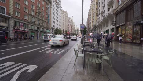 Madrid-Gran-Via-An-Einem-Regnerischen-Tag-Fußgänger,-Leere-Restauranttische-Und-Autos-In-Der-Belebten-Straße-Der-Spanischen-Stadt