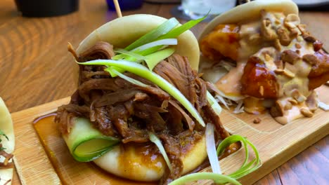 Three-delicious-hot-steam-bao-buns-with-pulled-pork,-coriander-and-crispy-onions,-duck-and-cucumber,-fried-chicken-and-peanuts,-traditional-Asian-food-at-a-restaurant,-4K-shot