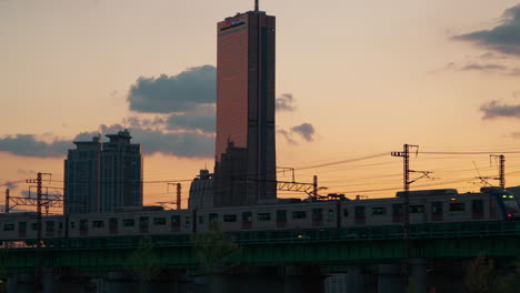 Seouler-U-Bahn-Fährt-Bei-Sonnenuntergang-Auf-Der-Hangang-Eisenbahnbrücke,-Gebäude-63-Vor-Farbenfrohem,-Orangefarbenem,-Atemberaubendem-Himmelshintergrund