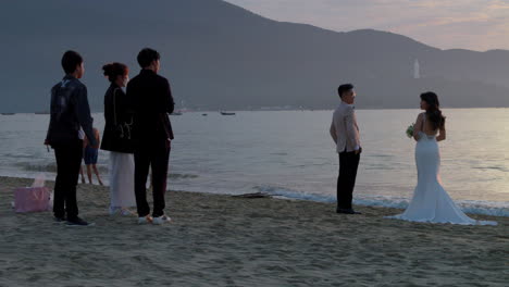 Wedding-photographer-poses-beautiful-asian-couple-at-sunset-as-ocean-waves-crash-on-beach