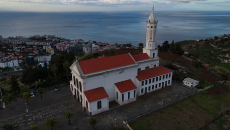 Iglesia-De-Sao-Martinho-En-Funchal,-Madeira:-Vista-Aérea-De-La-Hermosa-Iglesia-Y-Su-Torre-Blanca-Durante-La-Puesta-De-Sol,-Con-Vistas-A-La-Ciudad-Y-Al-Océano