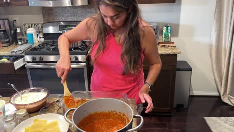Hispanic-young-woman-spreading-pasta-sauce-over-lasagna-noodles-in-glass-backing-dish,-in-home-kitchen