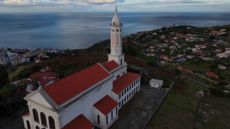 Aerial-view-over-the-Sao-Martinho-church-in-Funchal,-Madeira:-orbit-from-the-back-of-the-beautiful-church-during-sunset-and-overlooking-the-city-and-the-ocean
