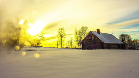Timelapse-shot-of-sun-setting-in-the-background-over-snow-covered-landscape-alongside-a-wooden-cottage-during-evening-time