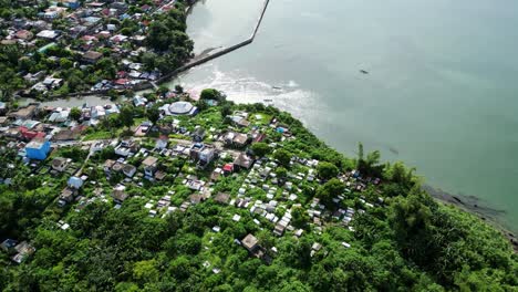 Aerial-pullback-view-of-tropical,-overgrown-cemetery-facing-stunning-ocean-bay-waters-and-local-village